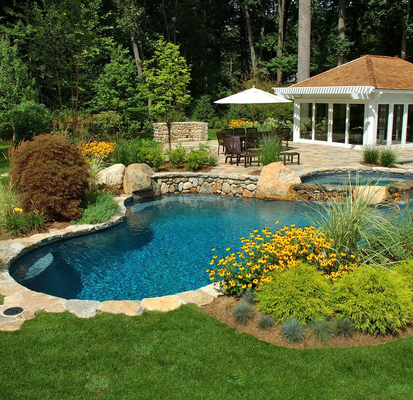 A inground backyard pool with hot tub, patio, pool house, and lush landscaping.