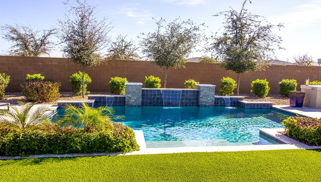 A backyard swimming pool with built-in waterfalls, bordered by shrubs and trees.