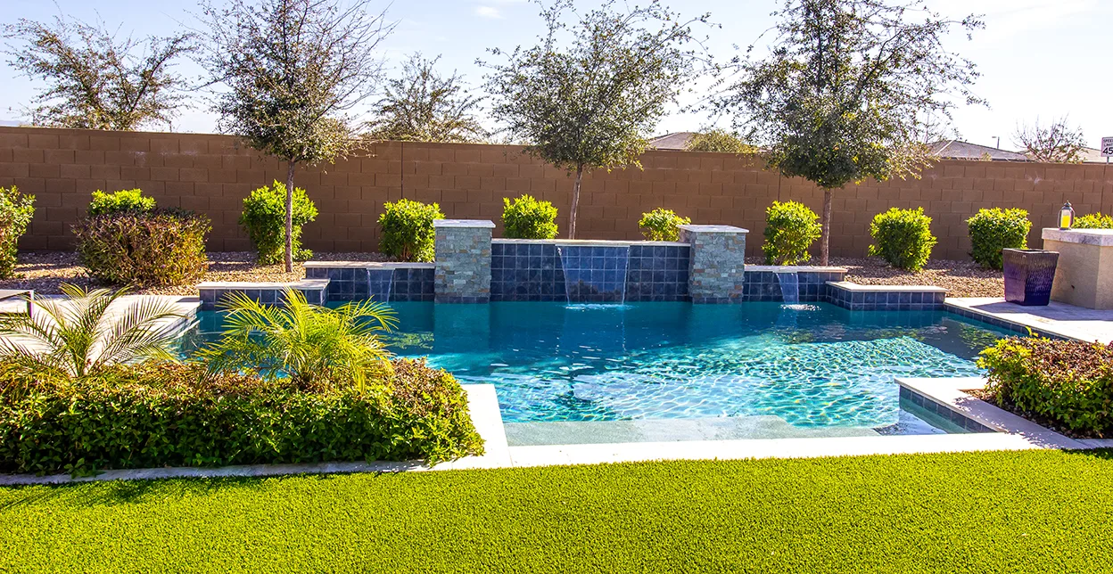 An inground swimming pool with built-in waterfalls, bordered by shrubs and trees.