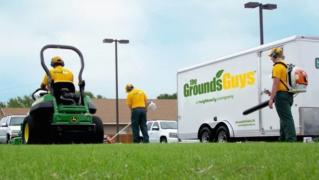 Three Grounds Guys service professionals performing lawn cleanup next to a branded company trailer.