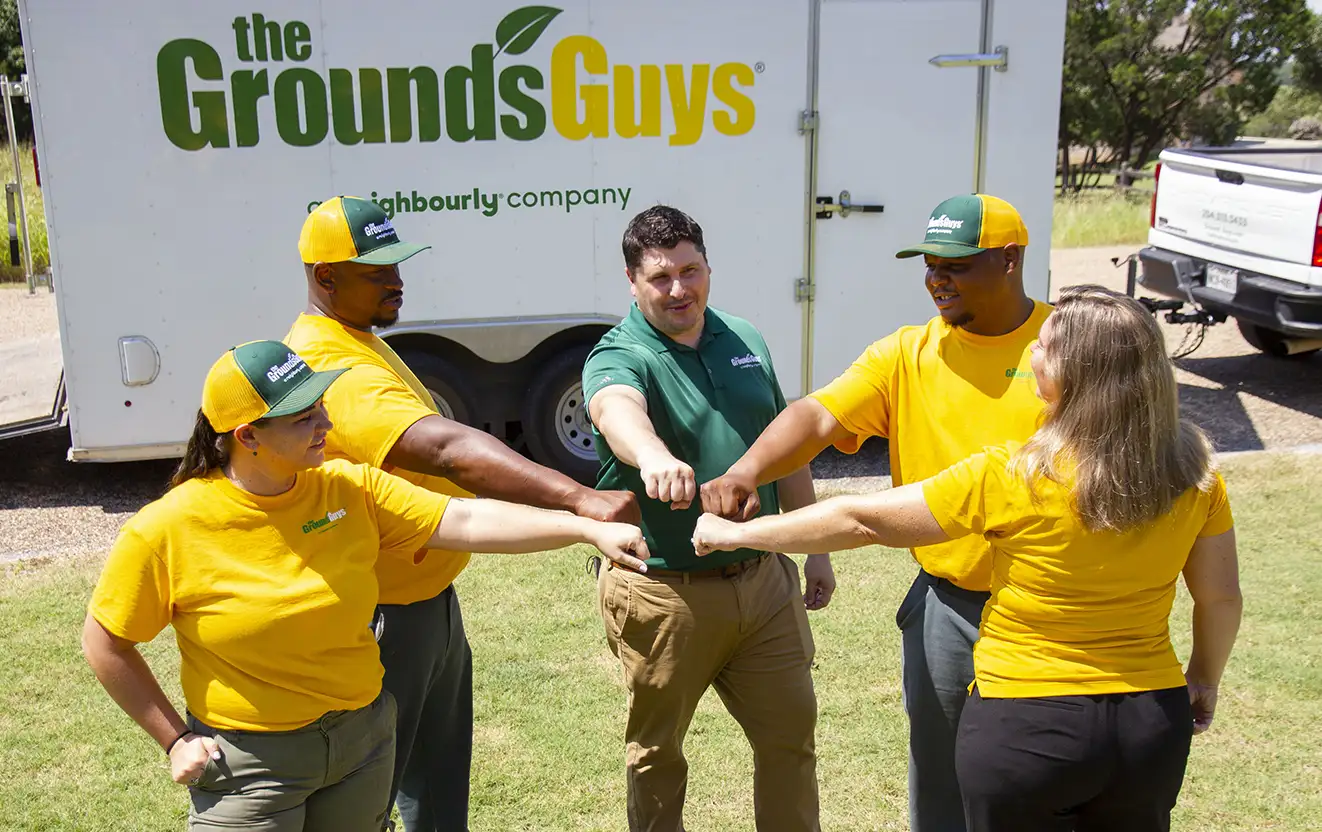 The Grounds Guys crew standing in a circle with their hands in the center, in front of a branded company trailer.